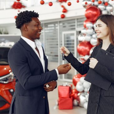 A woman working at a car dealership hands a man car keys as they stand in the dealership in front of a red car