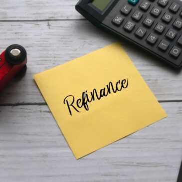 A yellow sticky note with "refinance" written on it appears on a desk next to a calculator and pen.