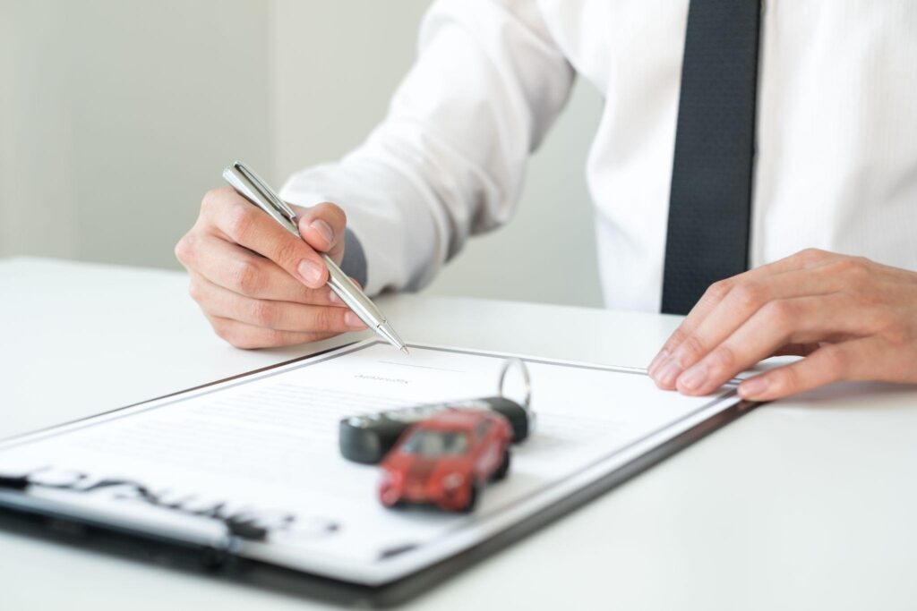 A mini car and keys appear on a clipboard as a man in a suit signs a document